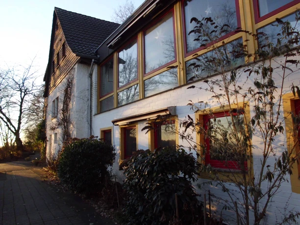 Gesundheitshaus-am-Wiehengebirge2.jpg Ein größeres Gebäude mit weißer Fassade, rotem Fensterrahmen, umgeben von Sträuchern im Herbstlicht.