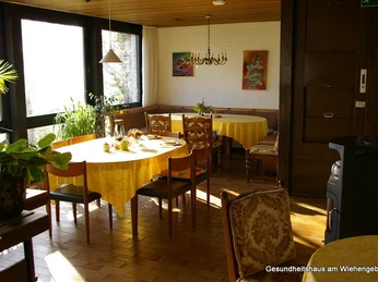 Heller Speiseraum mit gelb gedeckten Tischen, Stühlen und Pflanzen am Fenster. Kunstbilder an der Wand.