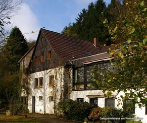 Zweistöckiges Gebäude mit Satteldach, umgeben von Bäumen. Weiße Fassade mit Holzverkleidung.