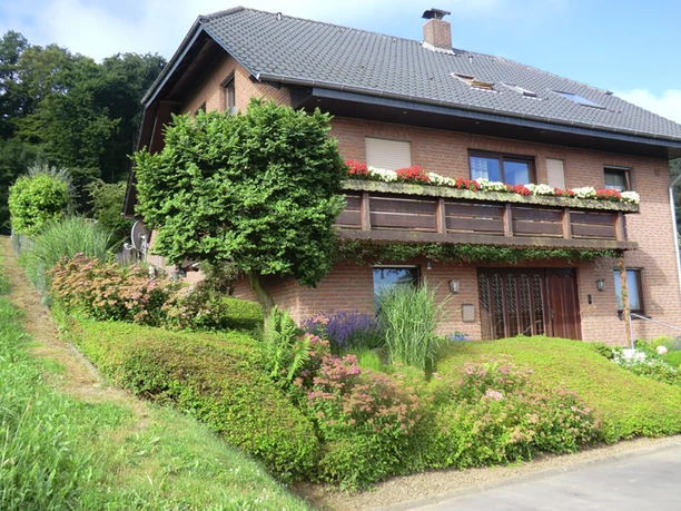 Am Huenenbrink-1.jpg Zweistöckiges Backsteinhaus mit begrüntem Vorgarten, Dachgauben und blühendem Blumenkasten am Balkon.