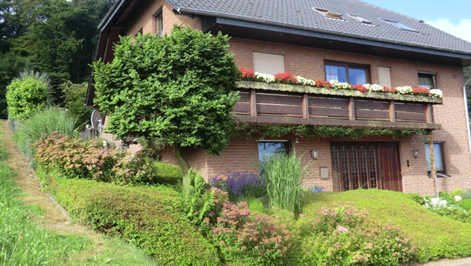 Am Huenenbrink-1.jpg Zweistöckiges Backsteinhaus mit begrüntem Vorgarten, Dachgauben und blühendem Blumenkasten am Balkon.