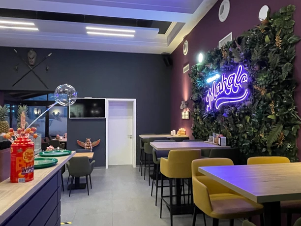 5.jpg Auf dem Bild ist die Inneneinrichtung des Restaurants Mahalo zu sehen. Rechts besteht ein Teil der Wand aus grünen Plastikpflanzen, an denen der leuchtende lilafarbene Schriftzug „Mahalo“ prangt. Auf der linken Seite stehen graue Tische und Stühle, in der Mitte des Bildes ist eine weiße Tür, und rechts befinden sich weitere Tische.