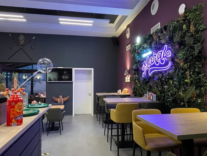 5.jpg Auf dem Bild ist die Inneneinrichtung des Restaurants Mahalo zu sehen. Rechts besteht ein Teil der Wand aus grünen Plastikpflanzen, an denen der leuchtende lilafarbene Schriftzug „Mahalo“ prangt. Auf der linken Seite stehen graue Tische und Stühle, in der Mitte des Bildes ist eine weiße Tür, und rechts befinden sich weitere Tische.