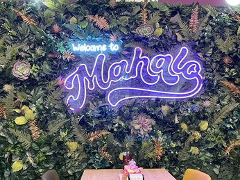 8.jpg Zu sehen ist der leuchtende lilane Schriftzug „Welcome to Mahalo“ auf einer Wand aus grünen Pflanzen.