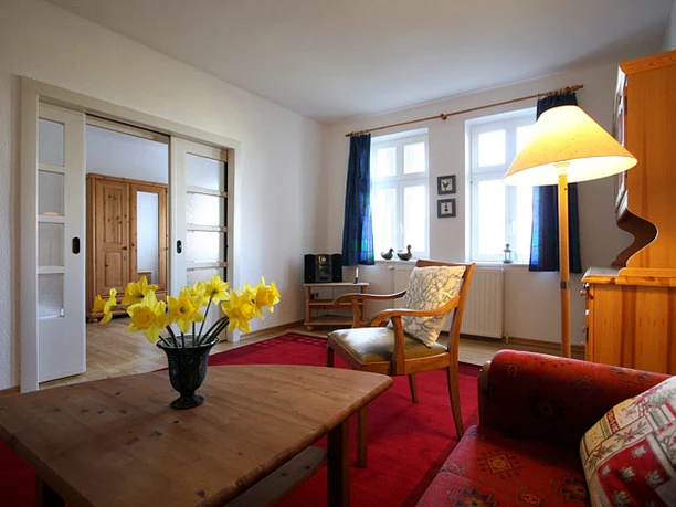 Hof Husemeyer Wohnzimmer mit Holzmöbeln, Teppich, roten Sofas und Vase mit gelben Blumen auf einem Tisch.