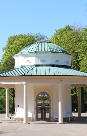 Runder Pavillon mit Säulen und grüner Kuppel in einem grünen Kurpark, umgeben von Bäumen.
