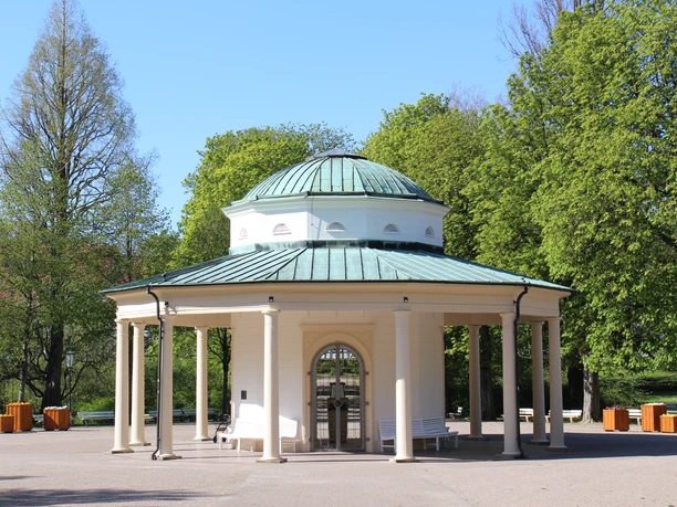 Runder Pavillon mit Säulen und grüner Kuppel in einem grünen Kurpark, umgeben von Bäumen.