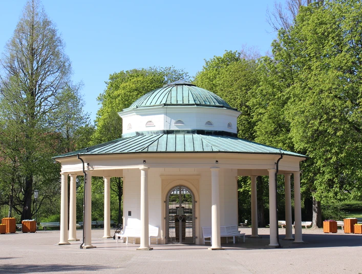 Brunnentempel im Historischen Kurpark Runder Pavillon mit Säulen und grüner Kuppel in einem grünen Kurpark, umgeben von Bäumen.