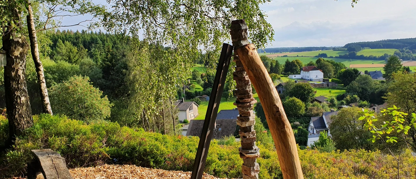 Ein Holzkunstwerk ragt auf einem bewaldeten Hügel empor, umgeben von grüner Landschaft.
