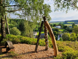 Dreiklang.jpg Ein Holzkunstwerk ragt auf einem bewaldeten Hügel empor, umgeben von grüner Landschaft.