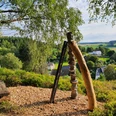 Dreiklang.jpg Ein Holzkunstwerk ragt auf einem bewaldeten Hügel empor, umgeben von grüner Landschaft.