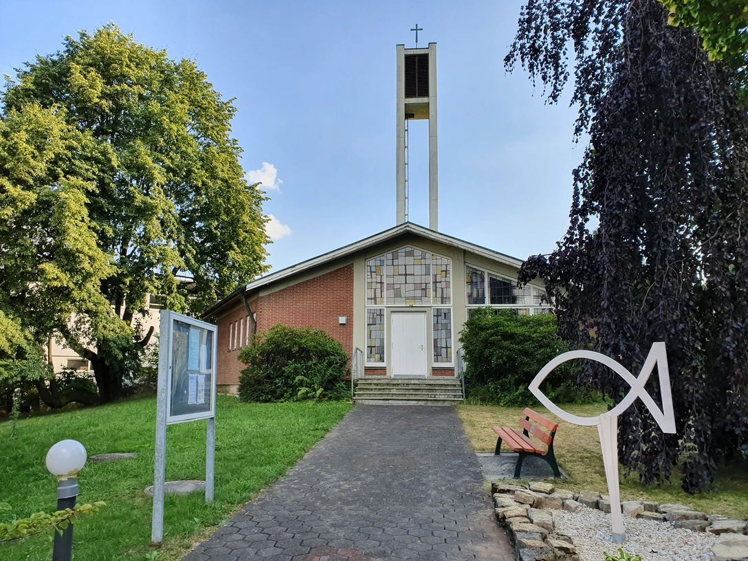 Evangelische Kirche mit hohem Glockenturm, gelegen in einer grünen Umgebung mit Parkbank und Gehweg.