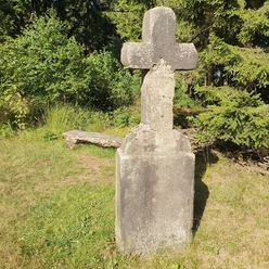 Feldromer Kreuz.jpg Altes Steinkreuz auf einer Wiese, umgeben von Bäumen und Pflanzen, mit sichtbarer Verwitterung.