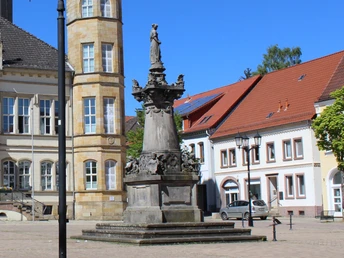 Steinernes Denkmal mit Figuren auf einem Sockel, umgeben von historischen Gebäuden und Bäumen.