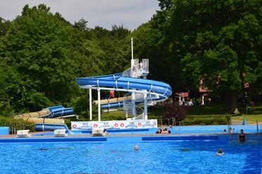 Das Bild zeigt ein Freibad mit einer großen Wasserrutsche, umgeben von grünen Bäumen und Schwimmern.