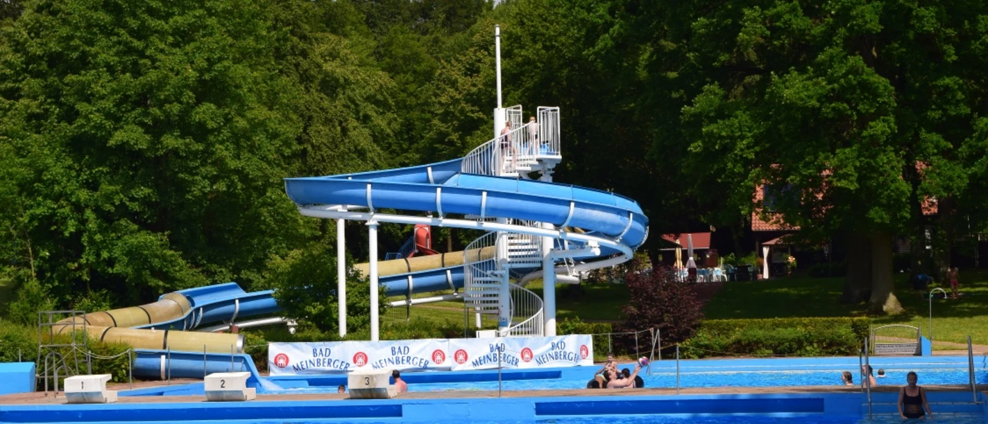 Freibad Bad Meinberg.png Das Bild zeigt ein Freibad mit einer großen Wasserrutsche, umgeben von grünen Bäumen und Schwimmern.