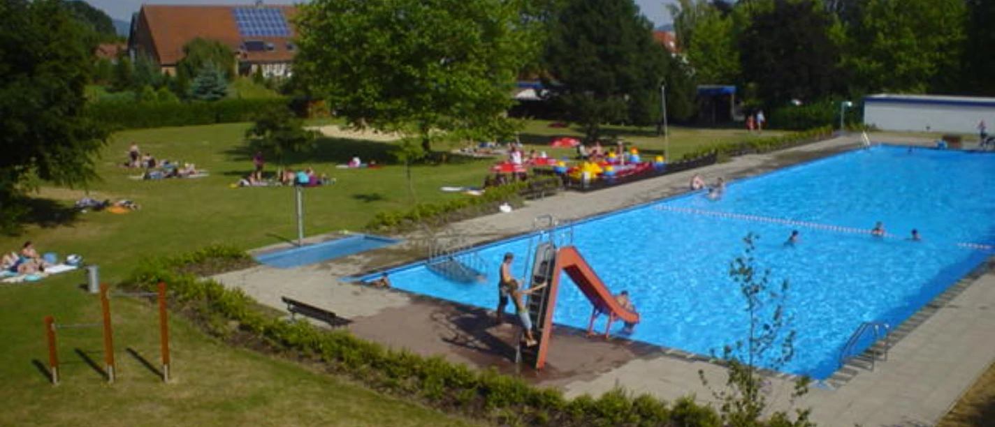 Freibad mit großem Schwimmbecken, Rutsche, Liegewiese und vielen Besuchern an einem sonnigen Tag.