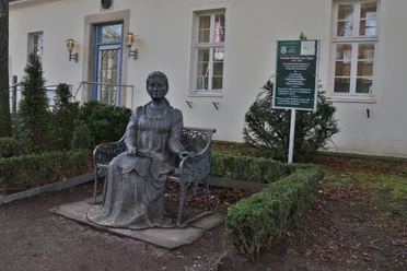 Bronzestatue der Fürstin Pauline mit Buch in der Hand, vor einem Gebäude mit Schautafel, umgeben von Büschen.