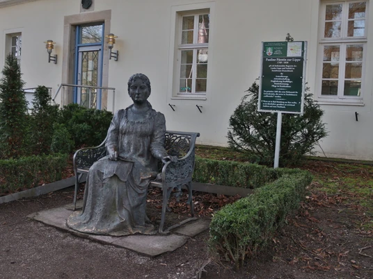 Fürstin Pauline Bronzestatue der Fürstin Pauline mit Buch in der Hand, vor einem Gebäude mit Schautafel, umgeben von Büschen.
