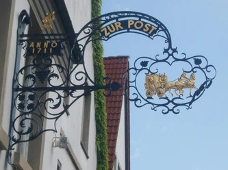 Schild zur Post.jpg Schwarzes, kunstvoll geschmiedetes Wirtshausschild "Zur Post" mit golderner Postkutsche und Pferden.