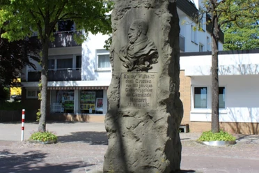 Kaiser-Wilhelm-Denkmal.png Steinernes Denkmal mit Kaiser Wilhelm-Büste, umgeben von Stadtgebäuden und Bäumen.