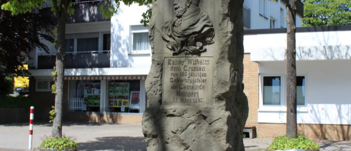 Kaiser-Wilhelm-Denkmal.png Steinernes Denkmal mit Kaiser Wilhelm-Büste, umgeben von Stadtgebäuden und Bäumen.