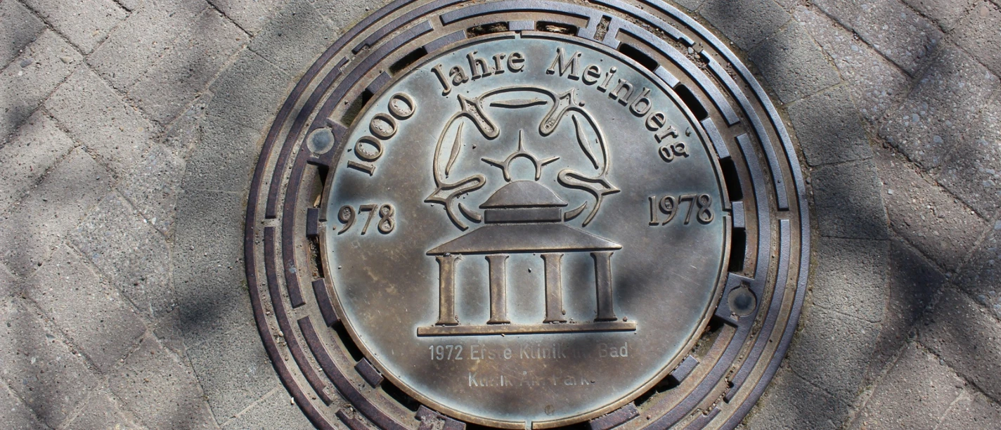 Kanaldeckel in Bad Meinberg, graviert mit "1000 Jahre Meinberg 1978" und einem stilisierten Kurhaus.