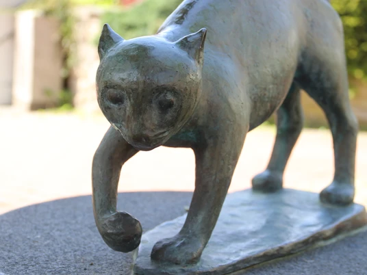 Bronzene Katzenfigur, die mit gehobener Pfote auf einem Sockel steht, mit sanftem, nachdenklichem Blick.