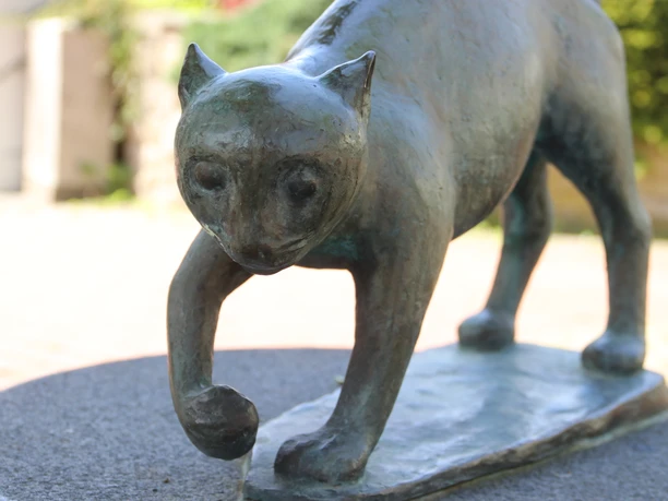 Bronzene Katzenfigur, die mit gehobener Pfote auf einem Sockel steht, mit sanftem, nachdenklichem Blick.