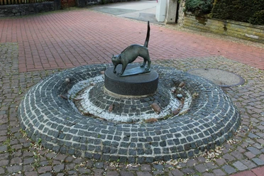 Bronzeskulptur einer Katze auf einem runden, erhöhten Sockel in einer gepflasterten Brunnenanlage.