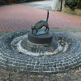 Katzenbrunnen Bronzeskulptur einer Katze auf einem runden, erhöhten Sockel in einer gepflasterten Brunnenanlage.