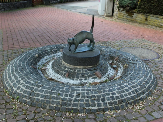 Katzenbrunnen Bronzeskulptur einer Katze auf einem runden, erhöhten Sockel in einer gepflasterten Brunnenanlage.