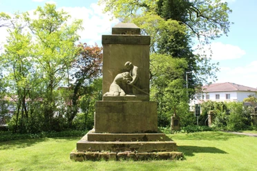 Denkmal aus Stein, das einen knienden Soldaten darstellt, umgeben von Bäumen auf grüner Wiese.