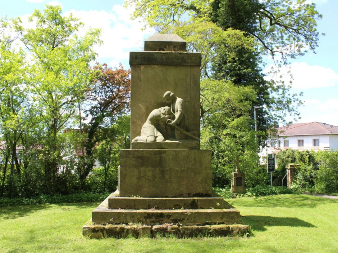 Denkmal aus Stein, das einen knienden Soldaten darstellt, umgeben von Bäumen auf grüner Wiese.