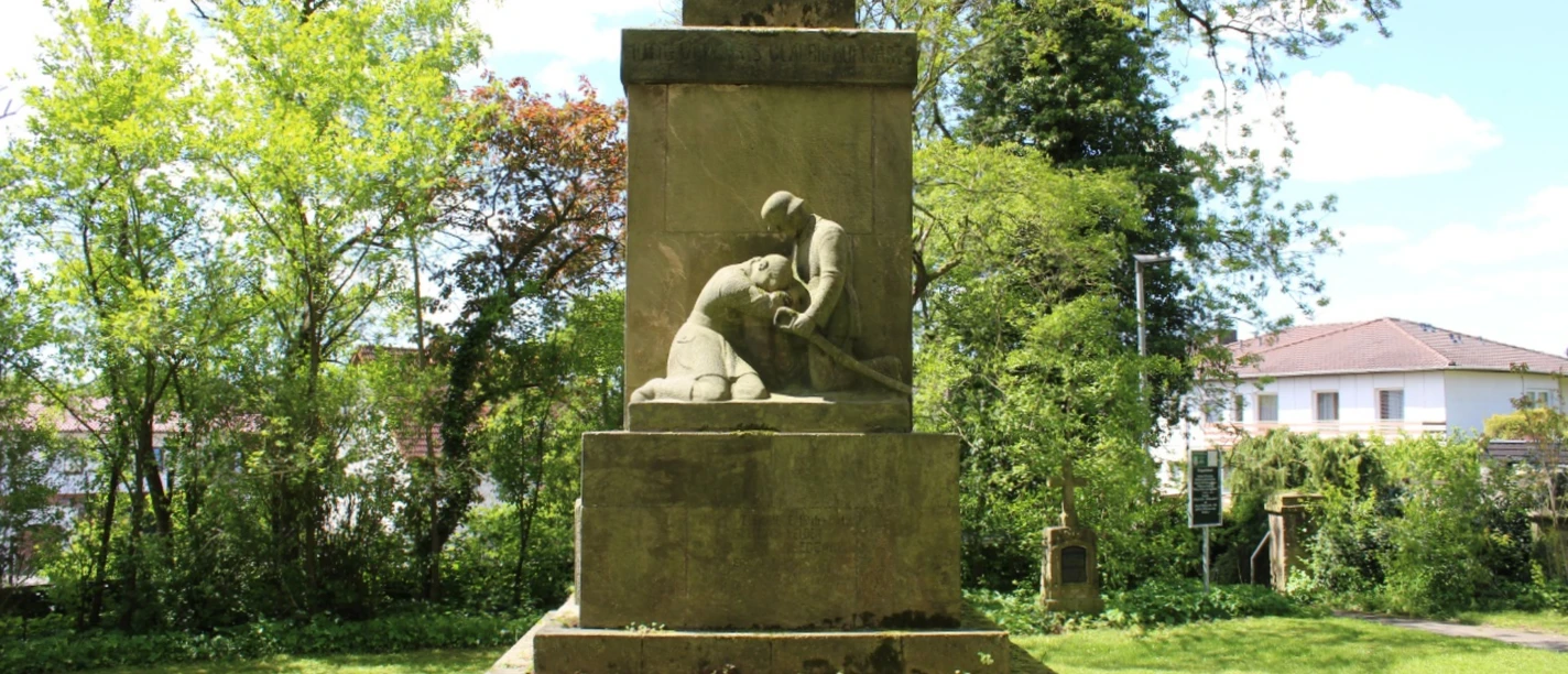Kriegerdenkmal.png Denkmal aus Stein, das einen knienden Soldaten darstellt, umgeben von Bäumen auf grüner Wiese.