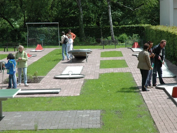 Menschen spielen auf einer Minigolfanlage mit Rasen und gepflasterten Wegen zwischen Bäumen und Büschen.
