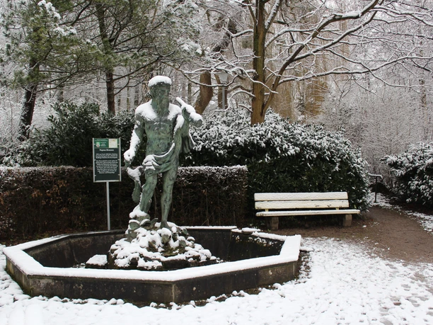 Verschneite Neptunstatue in einem ruhigen Garten, umgeben von schneebedeckten Büschen und Bäumen.