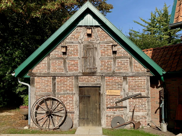 image00015.jpeg Altes Fachwerkhaus mit Holzreifen und Pflug an der Fassade unter blauem Himmel.