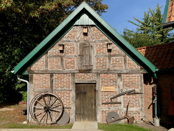 image00015.jpeg Altes Fachwerkhaus mit Holzreifen und Pflug an der Fassade unter blauem Himmel.