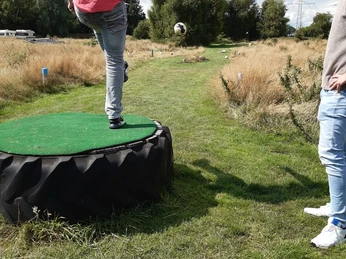 image00002.jpeg Auf dem Bild sieht man einen jungen Mann in Jeans und rotem T-Shirt, der auf einem Grasweg Fußball spielt. Er steht auf einer erhöhten Plattform aus Reifen, bereit, den Ball zu treten. Ein weiterer Mann schaut ihm zu. Im Hintergrund sind Bäume und ein grasbewachsener Pfad zu sehen, der in die Ferne führt.