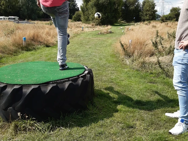 image00002.jpeg Auf dem Bild sieht man einen jungen Mann in Jeans und rotem T-Shirt, der auf einem Grasweg Fußball spielt. Er steht auf einer erhöhten Plattform aus Reifen, bereit, den Ball zu treten. Ein weiterer Mann schaut ihm zu. Im Hintergrund sind Bäume und ein grasbewachsener Pfad zu sehen, der in die Ferne führt.