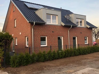 Backsteinhaus mit blauen Dachziegeln und Dachfenstern, umgeben von jungen Heckenpflanzen.