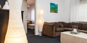 Ferienwohnung Berger 1.jpg Gemütliches Wohnzimmer mit braunem Sofa, hellem Teppich, Stehleuchten und Tür zu angrenzendem Raum.