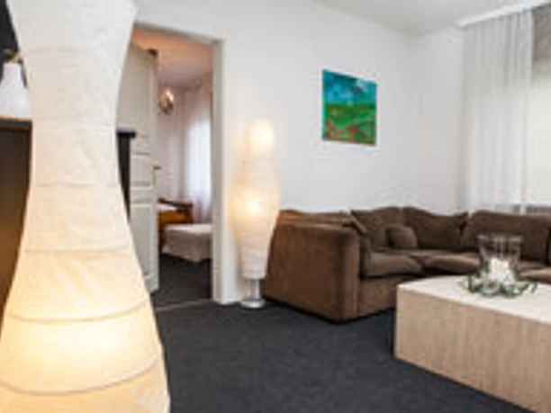 Ferienwohnung Berger 1.jpg Gemütliches Wohnzimmer mit braunem Sofa, hellem Teppich, Stehleuchten und Tür zu angrenzendem Raum.