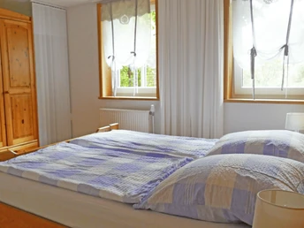 Doppelbett mit blau-weiß karierter Bettwäsche, Holzmöbel und Fenster mit lichtdurchlässigen Vorhängen.