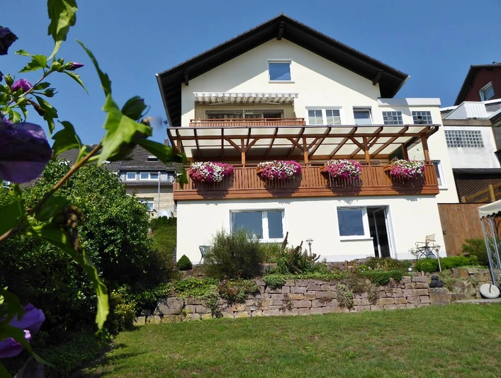 Ansicht.JPG Weißes Haus mit Balkon aus Holz und bunt blühenden Blumen, gepflegter Garten im Vordergrund
