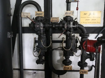 Historische Pumpenanlage mit Rohren und Schildern, darunter "Sauen", "Kessel", "Drücken", 80-Jahre-Tafel.