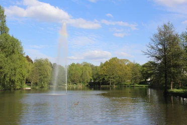 Ein ruhiger See mit einer hohen Wasserfontäne, umgeben von üppigem Grün und vereinzelten Enten.