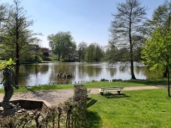 Blick von der Neptunstatue Eine ruhige Parkszene mit einem Teich im Hintergrund, eingerahmt von grünen Bäumen und Wiesen.