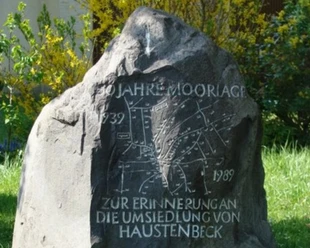 Gedenkstein mit eingravierter Landkarte und der Aufschrift "50 Jahre Moorlage" zwischen Pflanzen.
